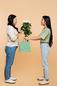 full length of happy asian daughter giving paper bag and flowers to mother on beige