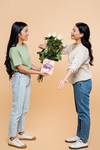 happy asian daughter congratulating mother with greeting card and flowers on beige