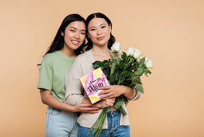 happy asian woman hugging mother with flowers and greeting card isolated on beige