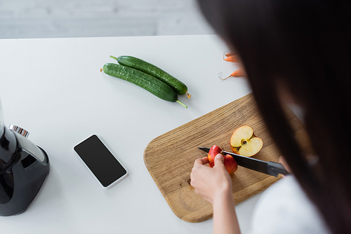 overhead view of blurred woman cutting apple near smartphone with blank screen and fresh cucumbers