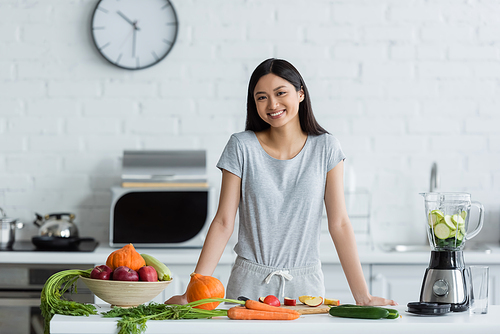 cheerful asian woman  near electric blender and fresh vegetables in kitchen