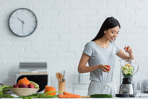 happy asian woman adding apple into shaker with sliced zucchini near fresh vegetables in kitchen