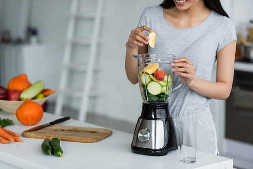 partial view of smiling woman adding apple into electric blender with sliced zucchini