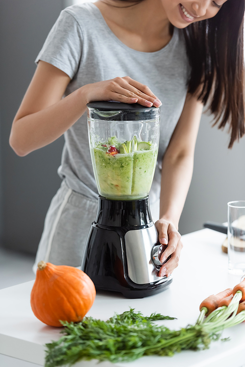 partial view of smiling asian woman preparing smoothie near raw pumpkin and carrots