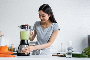 cheerful asian woman preparing vegetable smoothie in electric shaker