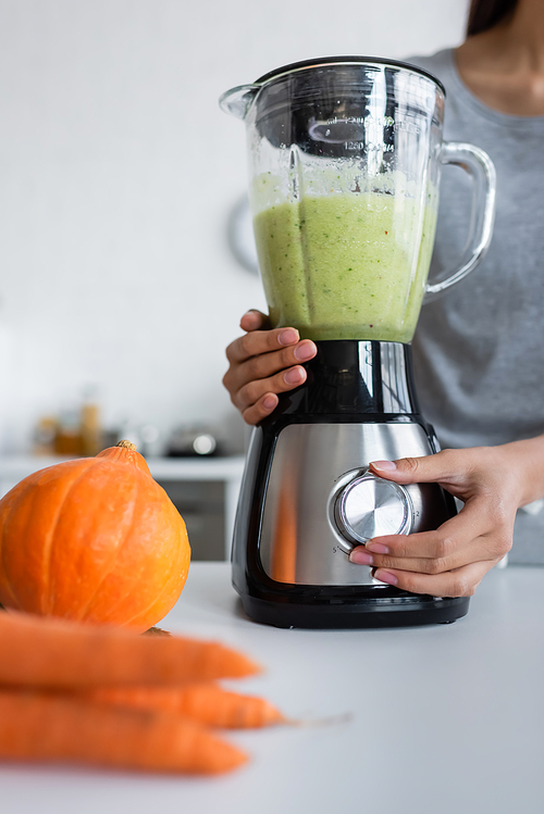 cropped view of woman preparing vegetable smoothie near pumpkin and blurred carrots