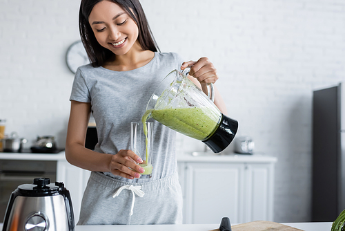 happy asian woman pouring fresh smoothie into glass in kitchen