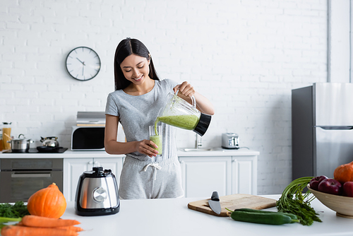 smiling asian woman pouring homemade smoothie near fresh vegetables in kitchen