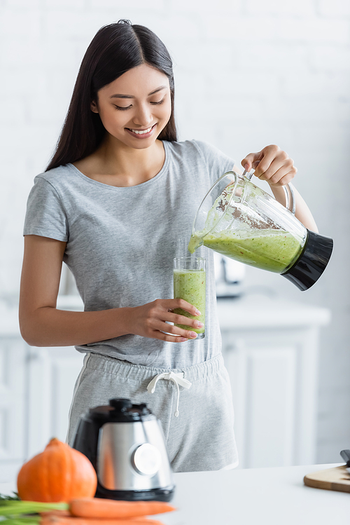 cheerful asian woman pouring homemade smoothie into glass near blurred vegetables in kitchen