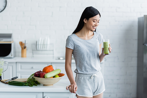happy asian woman standing with glass of homemade smoothie near bowl with fresh vegetables and apples