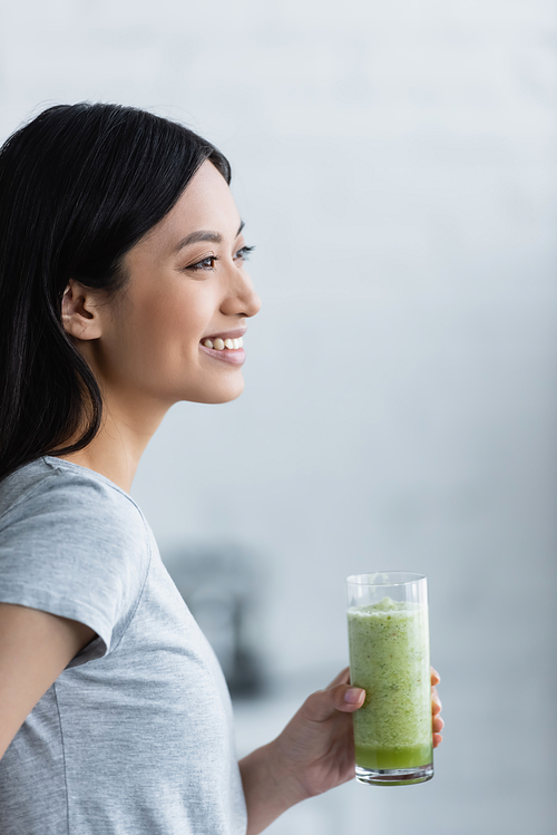 young asian woman smiling while holding glass of fresh smoothie at home