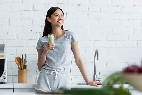 happy asian woman holding glass of fresh smoothie while standing in kitchen