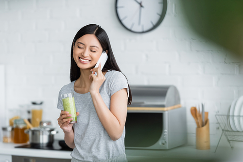 smiling asian woman with glass of fresh smoothie talking on cellphone in kitchen