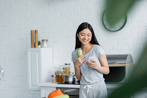 cheerful asian woman with glass of smoothie messaging on cellphone on blurred foreground