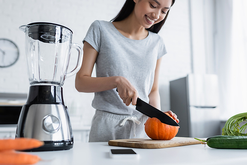 smiling asian woman cutting pumpkin near electric blender, smartphone and fresh vegetables