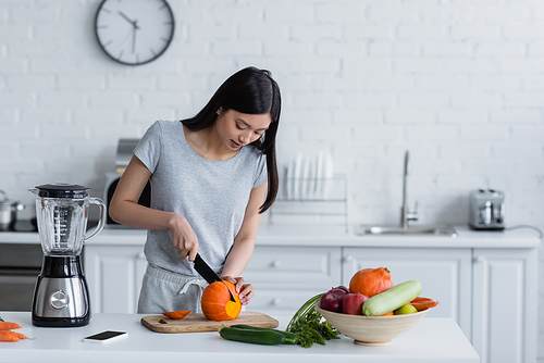 young asian woman cutting pumpkin near vegetables, electric blender and smartphone