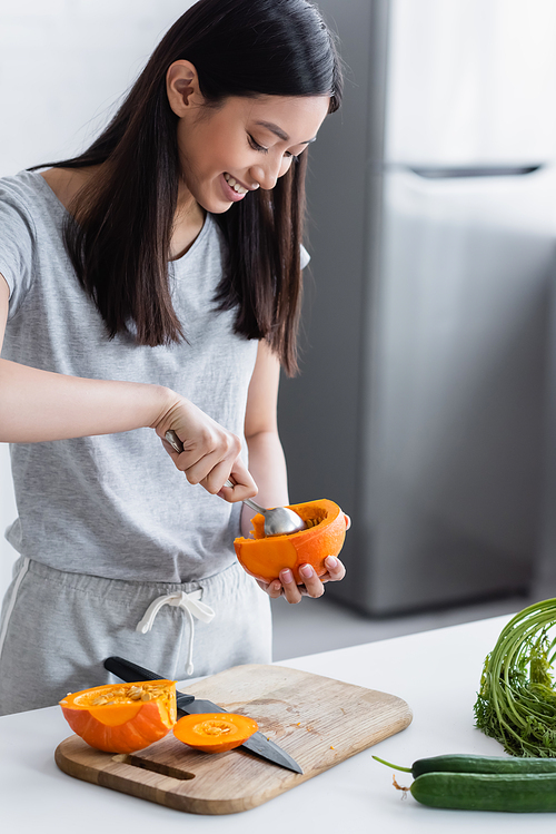 smiling asian woman cleaning out ripe pumpkin near fresh cucumbers on table