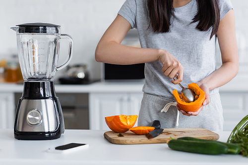 partial view of woman cutting out pumpkin with spoon near electric shaker, smartphone and blurred cucumbers