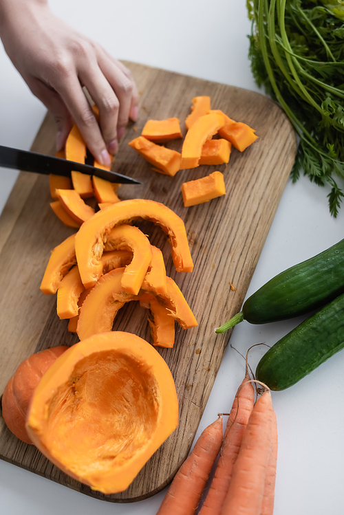 partial view of woman cutting ripe pumpkin on chopping board near fresh carrots and cucumbers