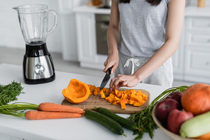 cropped view of woman cutting ripe pumpkin near fresh vegetables and electric blender