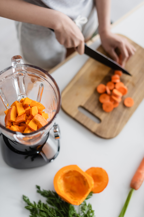 partial view of blurred woman cutting carrot near blender with pumpkin