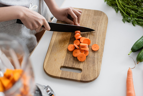 partial view of woman cutting raw carrot near fresh cucumbers on kitchen table