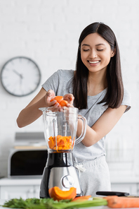 cheerful asian woman adding sliced carrot into electric blender with cut pumpkin