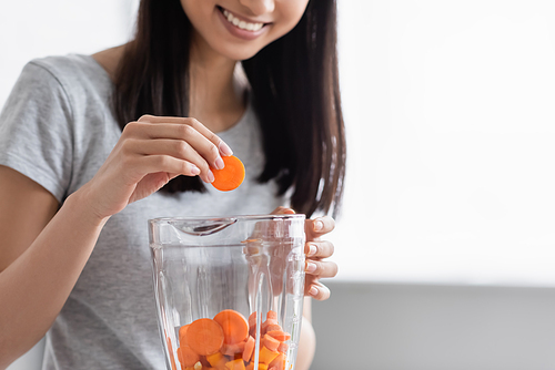 cropped view of smiling asian woman adding carrot into blender with cut pumpkin