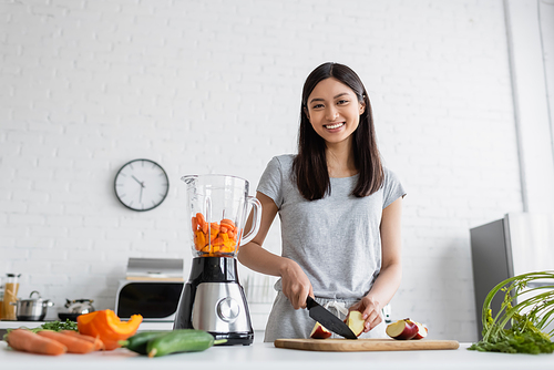 cheerful asian woman cutting apple near electric blender and fresh vegetables in kitchen