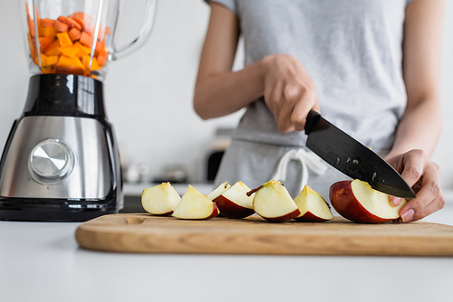 cropped view of blurred woman cutting apple on chopping board near electric shaker