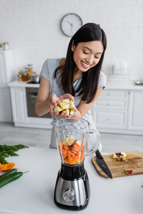 joyful asian woman adding cut apple into electric blender with raw vegetables
