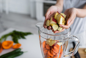 cropped view of woman adding apples into jug of blender with cut carrots