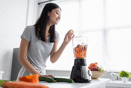 smiling asian woman adding apple into blender with fresh vegetables in kitchen