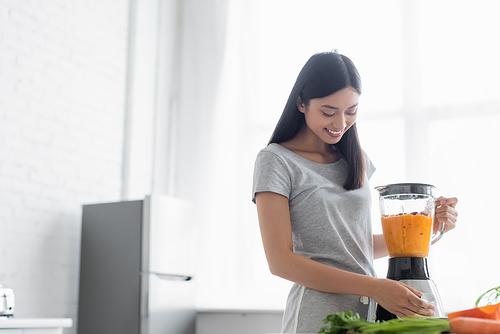cheerful asian woman preparing vegetable smoothie for breakfast in kitchen