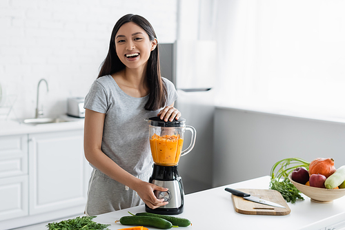 cheerful asian woman smiling at camera while preparing fresh vegetable smoothie