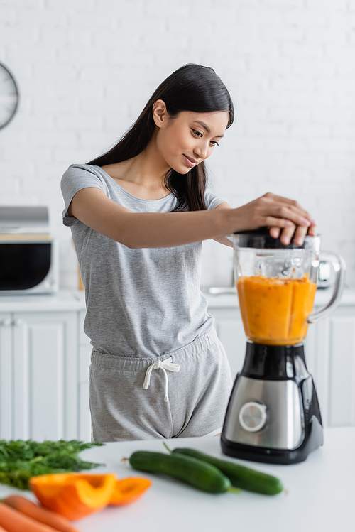 young asian woman preparing fresh smoothie in blender near blurred vegetables on table