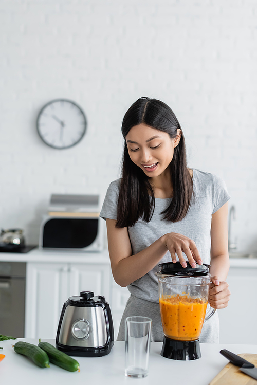 happy asian woman preparing smoothie in electric shaker near fresh cucumbers