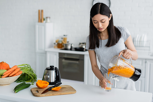 young asian woman pouring fresh smoothie near fresh vegetables and chopping board on kitchen table