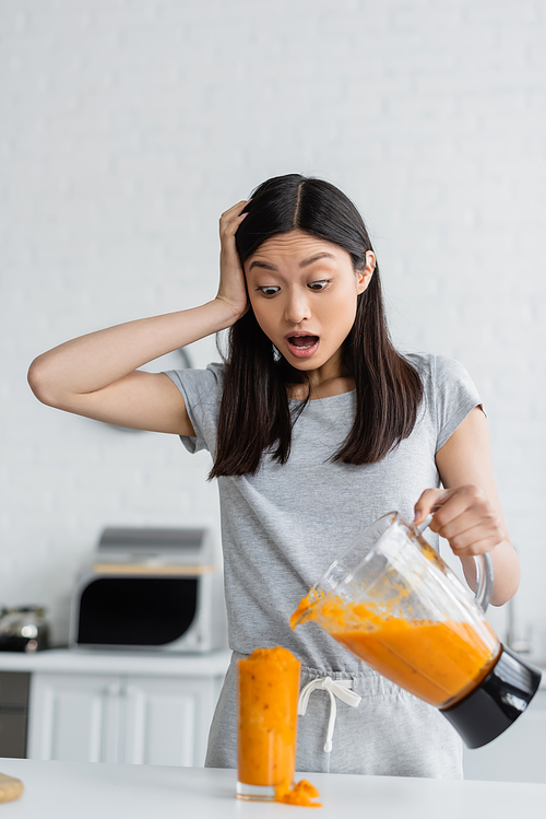 shocked asian woman touching head near overflowing glass of smoothie in kitchen