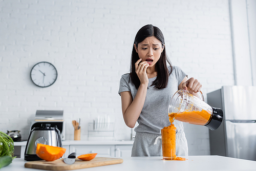 worried asian woman pouring smoothie into overflowing glass near cut pumpkin on chopping board