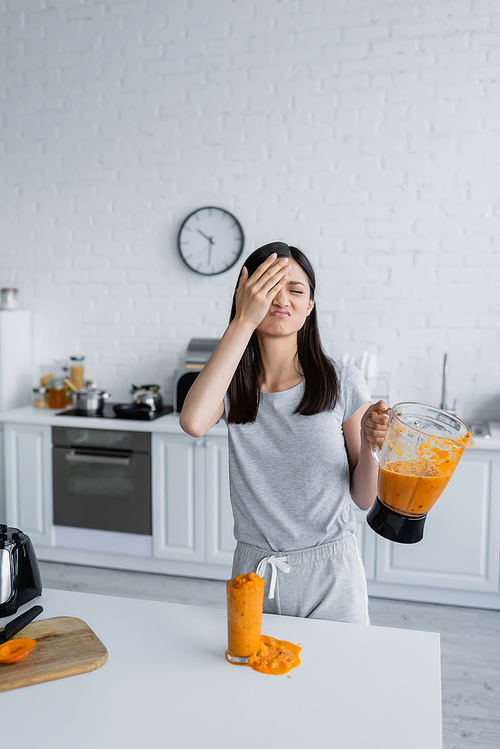 displeased asian woman covering face with hand while holding jar of smoothie near overflowing glass