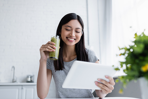 smiling asian woman looking at digital tablet while holding glass of tasty smoothie
