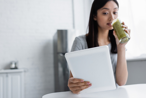 blurred asian woman drinking homemade smoothie while using digital tablet