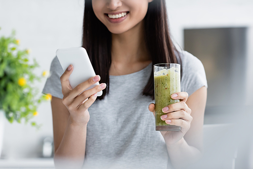 partial view of brunette woman with glass of smoothie messaging on smartphone on blurred foreground