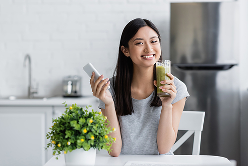 happy asian woman holding cellphone and glass of homemade smoothie near blurred potted plant