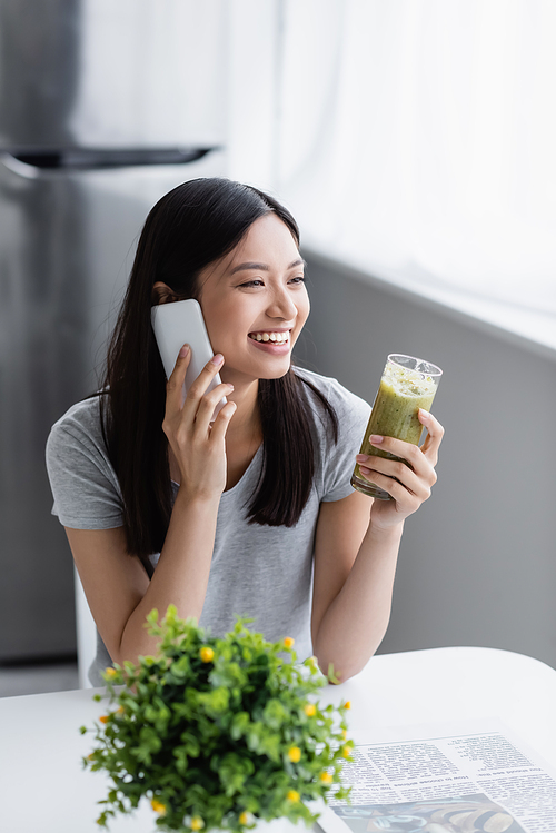 joyful asian woman with glass of fresh smoothie talking on mobile phone near blurred plant