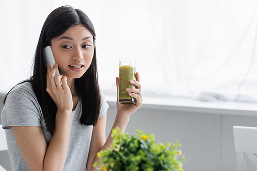 young asian woman calling on mobile phone while holding glass of smoothie near blurred plant