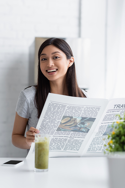 cheerful asian woman smiling at camera while standing with newspaper near blurred glass of smoothie
