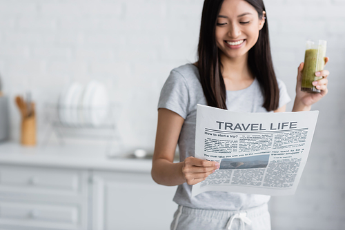 blurred asian woman with glass of fresh smoothie reading travel life newspaper in kitchen