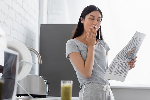 sleepy asian girl yawning while reading morning newspaper in kitchen